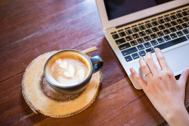 İşletme eliyle sıcak latte sanat kahvesi. Kadınlar kafedeki ahşap masada dizüstü bilgisayar kullanıyor. - Fotoğraf, Görsel