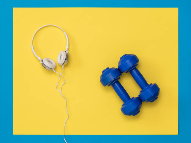 Kék súlyzók és fehér fülhallgatók sárga és kék háttérrel. - Fotó, kép