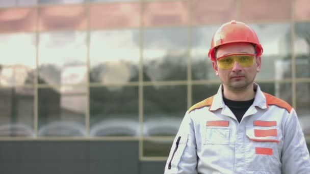 Portrait of an engineer or builder. A man (worker, engineer) in overalls, in a protective helmet and glasses. Behind him is a modern glass building. The camera moves from left to right. - Πλάνα, βίντεο