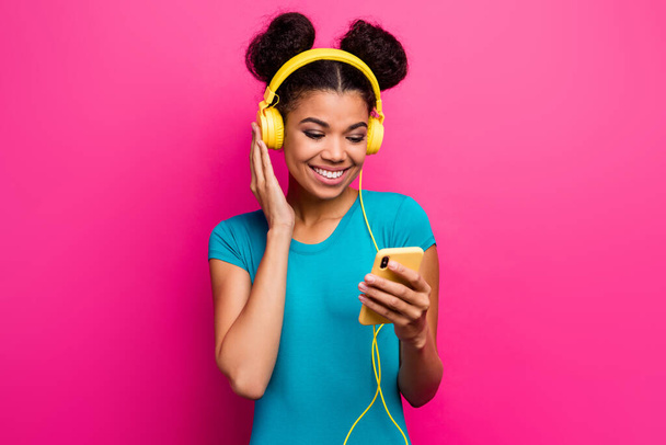 Photo of cheerful funny dark skin lady hold telephone hands choosing cool song to turn on use modern earphones wear blue casual t-shirt isolated bright pink color background - Foto, Imagem