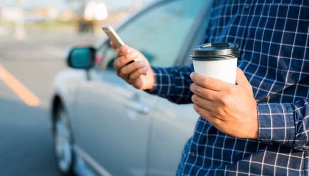 Close up male hand holding coffee paper cup and using smartphone near a car - Photo, Image