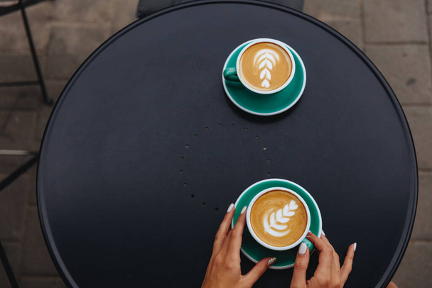 Top view of two turquoise-coloured cups of hot coffee with beautiful latte art in form of heart on black table outdoors. Young lady with trendy manicure holding one mug with morning drink in hands - Photo, Image