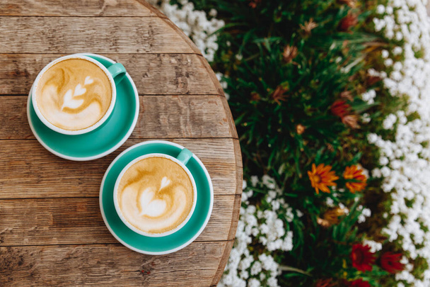 Cappuccino de aroma caliente con patrón de corazón hecho de espuma de leche en tazas de cerámica verde en mesa de madera redonda con hermosas flores que crecen cerca al aire libre. Cómoda terraza de verano para tomar un café
 - Foto, Imagen