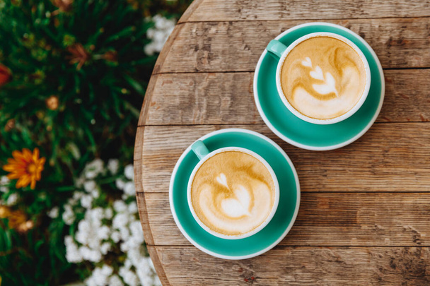 Draufsicht auf morgendlichen Aroma-Cappuccino mit Herzmuster in hellblauen Tassen mit Untertassen auf Holztisch mit Blumensträuchern in der Nähe. Sommerzeit auf der gemütlichen Café-Terrasse. - Foto, Bild