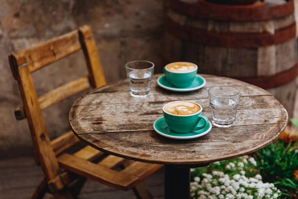 Bella terrazza caffè con sedia in legno e tavolo e verde tazze di ceramica di gustoso cappuccino aroma con schiuma di latte a forma di cuore. Mattina bevanda calda servita con bicchiere d'acqua
. - Foto, immagini