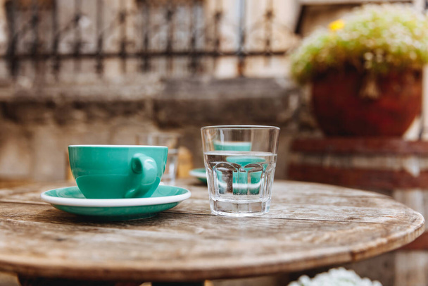 Grüne Keramiktasse Cappuccino-Kaffee auf einem runden Holztisch mit einem Glas Wasser auf der Sommerterrasse. Genuss von heißen Aromagetränken an frischer Luft. - Foto, Bild