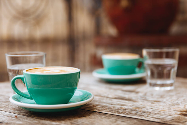 Δύο τυρκουάζ φλιτζάνια φρεσκοψημένου αρωματικού καφέ με πλούσιο γάλα σερβιρισμένο με ποτήρια νερό σε στρογγυλό ξύλινο τραπέζι στο καφέ. Vintage αποτέλεσμα και ζεστή ατμόσφαιρα - Φωτογραφία, εικόνα