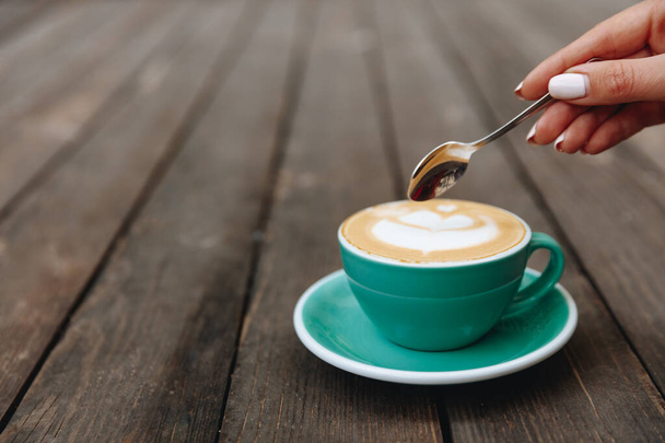 Female hand with white manicure holding spoon over hot fresh aroma cappuccino with latte art in light blue mug. Young beautiful girl is going to enjoy morning tasty beverage - Photo, Image