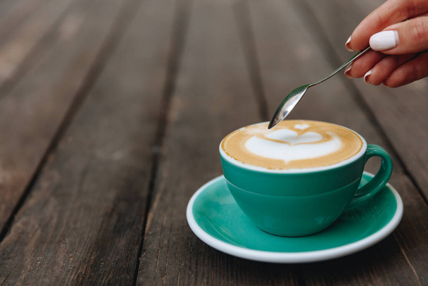  turquoise - coloured cup of freshly brewed coffee with lush milk foam on wooden background. Morning hot beverage served on saucer with tea spoon that holding woman's hand - Photo, Image