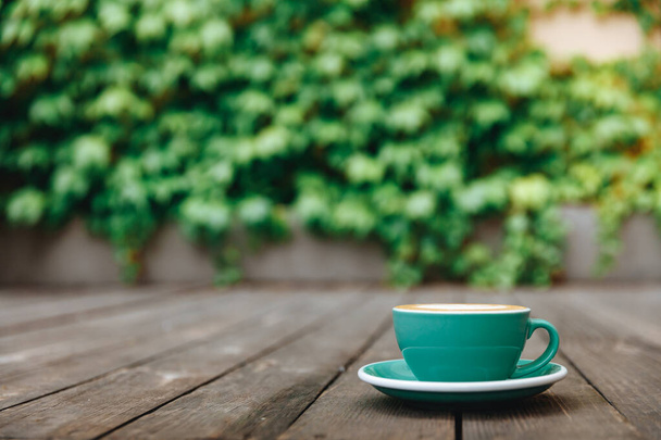 Freshly brewed coffee in turquoise-coloured cup with latte art on wooden table with blur background of green leaves. Start the day with tasty hot beverage - Photo, Image