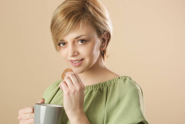 Studio shot of happy young woman holding cup of coffee and eating chocolate chip cookie - Foto, afbeelding