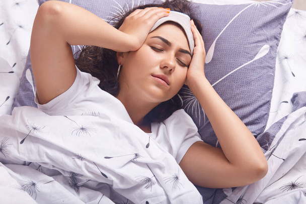 Indoor shot of brunette lady with blindfold on her forehead, lying with closed eyes and touching her temples, suffering from headache in morning, female staying in bed because of illness. Health care. - Photo, Image