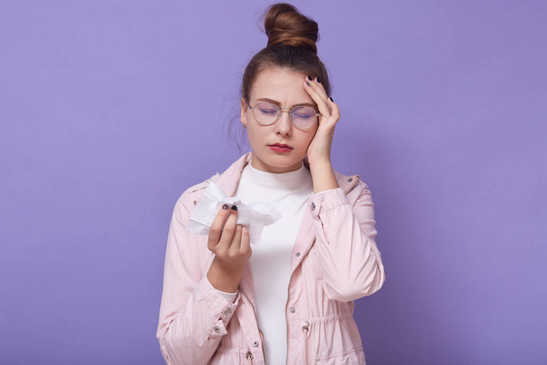 Horizontal shot of unhappy ill female closing eyes, putting fingers on forehead, holding handkerchief, feeling unwell, having running nose, standing isolated over lilac background. Health concept. - Photo, image