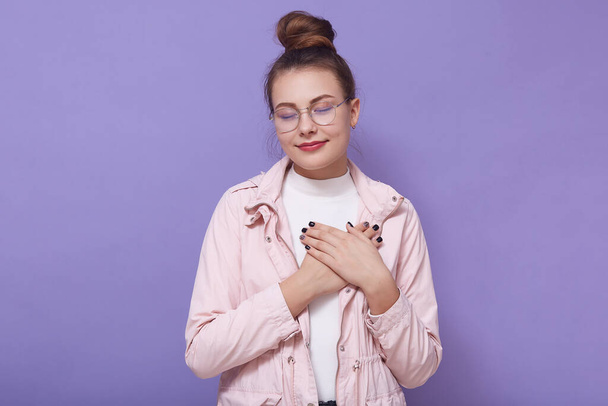 Indoor shot of pretty woman with closed eyes, keeping her hands on chest, being thankful, posing isolated over lilac background, adorable grateful female with hair bun and wearing jacket and eyewear. - Φωτογραφία, εικόνα