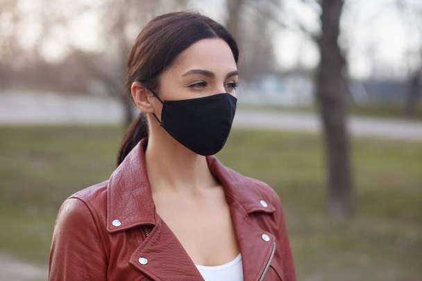 Outdoor shot of beautiful brunette woman wearing leather jacket and black protective mask, looking in distance, protecting herself from dangerous corona virus, walking in open air. Covid 19 concept. - Foto, Bild
