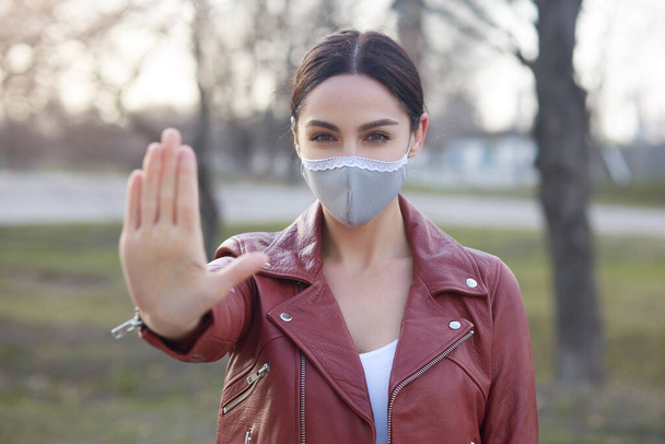 Outdoor shot of young adorable dark haired female posing in street and showing stop gesture with her palm, woman wearing protective mask, preventing fast spreading corona virus. Covid 19 concept. - Foto, Bild