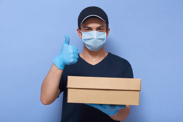 Closeup portrait of young guy working as courier or delivery man, wearing casual clothes and protective mask and gloves, holding carton box for customer and showing thumb up against blue wall. - 写真・画像