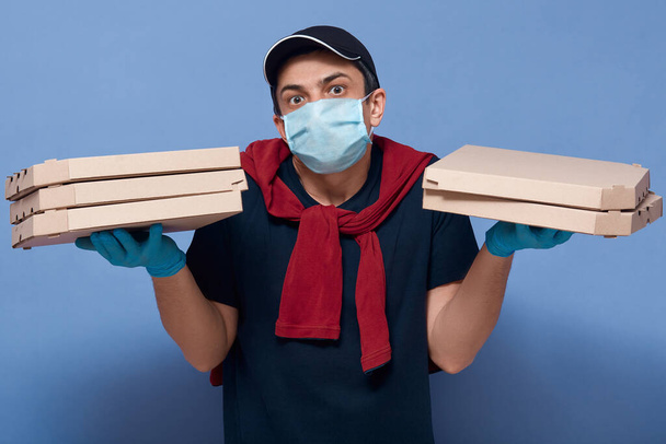 Indoor shot of confused surprised courier man holding carton boxes in both hands, having tired facial expression, wearing protective mask and gloves for protection. Quarantine and delivery concept. - Fotoğraf, Görsel