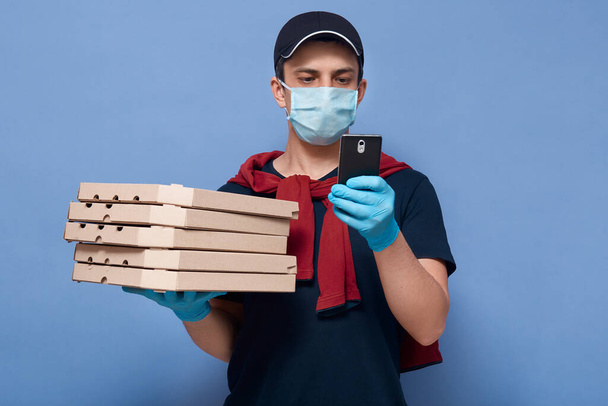 Closeup portrait of guy who works as online pizzeria employee, delivery man holding stack of pizza's boxes and smart phone in hands, tries to find address of customer, side job during quarantine. - Photo, Image