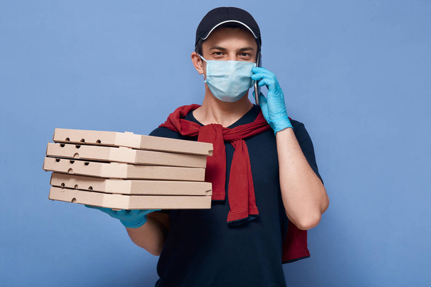 Picture of smiling delivery man talking to his client via smart phone, courier holding pizza's boxes and looking directly at camera, online order, special quarantine rules, male wears mask and gloves. - Φωτογραφία, εικόνα