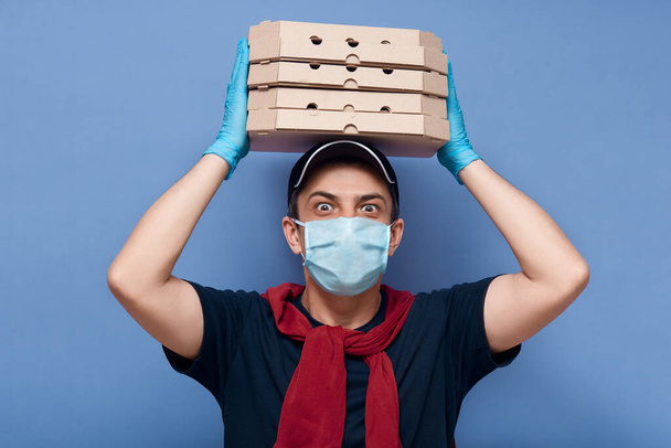 Studio shot of surprised delivery man dresses casual outfit, mask and latex gloves, holding stack of pizza's boxes above his head, has big eyes, looks shocked, delivers online order to customer. - Foto, Imagen