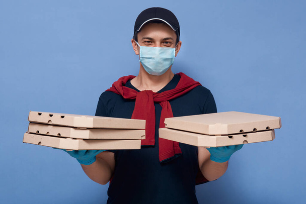 Studio shot of young handsome delivery man with carton pizza boxes in both hands, courier dresses casual outfit and protective mask and gloves isolated over blue background. Online ordering concept. - Photo, Image
