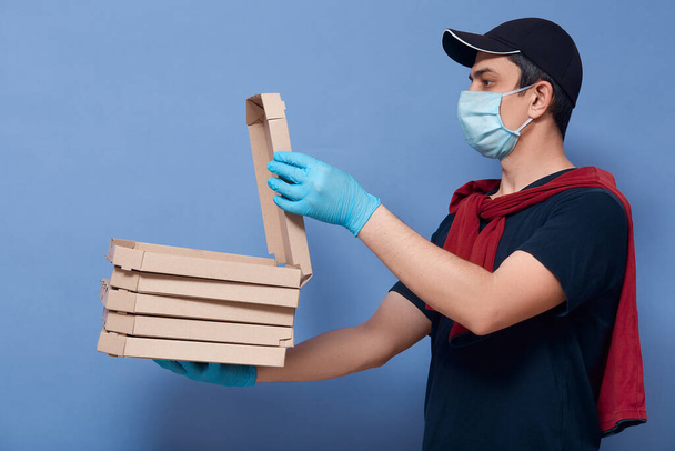 Profile of courier wearing t shirt, sweater over shoulders, cap, protective mask and latex gloves, delivery man opens box with order and shoving it to customer, posing isolated on blue background. , - Photo, Image