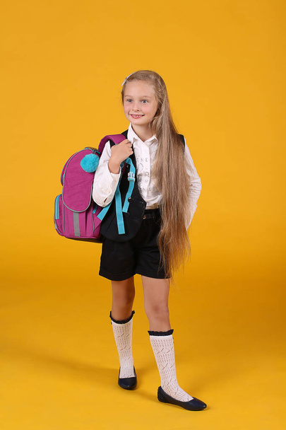 Birinci sınıf öğrencisi bir kızın portresi, okul üniforması içinde, sarı bir arka planda uzun dalgalı saçlı, sırt çantalı. - Fotoğraf, Görsel