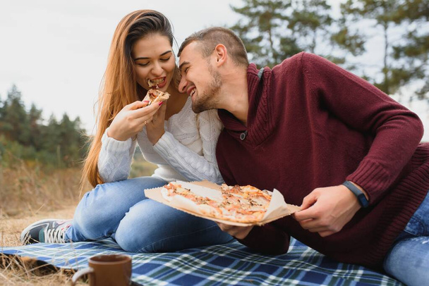 Young couple sitting on park bench and eating pizza.Fast food.Woman and man having picnic at sunset. Guys relaxing and enjoying food. Lifestyle - Фото, изображение