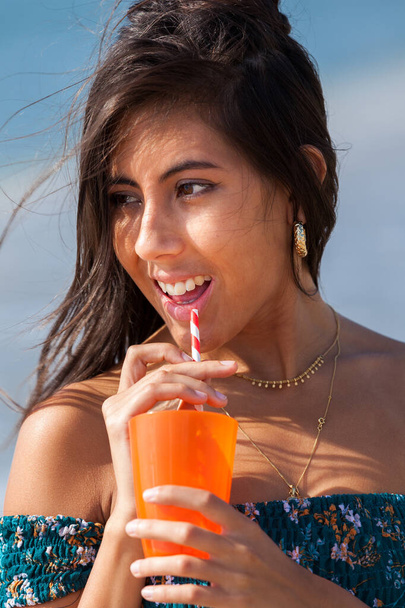 Portrait of a beautiful brunette drinking cocktail while standing on beach. Summer vacation concept. Happy girl enjoying freedom - Photo, Image
