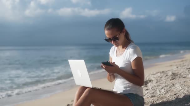 サングラスのフリーランサーはビーチでの作品のためのラップトップとスマートフォンを使用しています - 映像、動画
