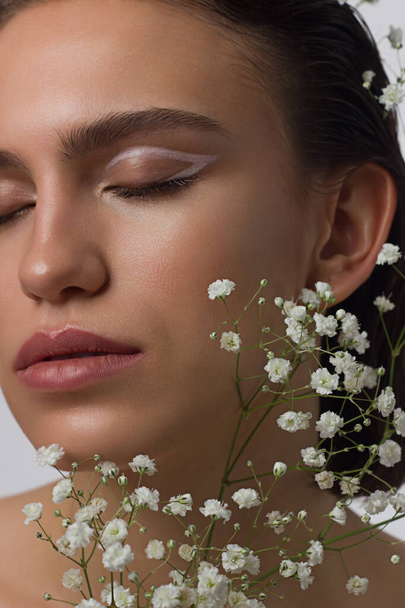 Close up portrait of a face with beautiful skin and nude makeup, eyeliner and long eyelashes, puffy natural lips and white flowers. Fashion photography. Cosmetology and spa. Freshness of spring - Foto, Imagem