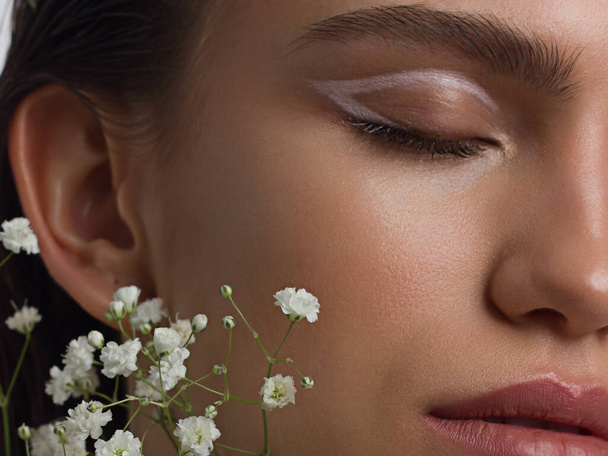 Close up half portrait of a face with beautiful skin and nude makeup, eyeliner and long eyelashes, puffy natural lips and white flowers. Fashion photography. Cosmetology and spa. Freshness of spring - Foto, immagini