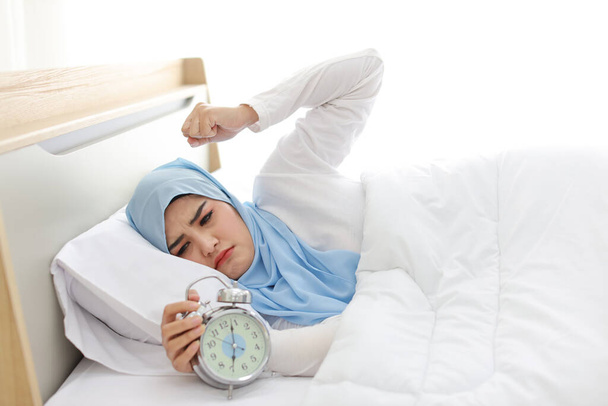 ベッドの上に寝そべって白い寝姿のアジア系イスラム教徒の女性が寝そべって、目覚まし時計の指輪を失って目を覚ます。サイドビューポートレート若い女の子は枕の上で頭を下げながら - 写真・画像