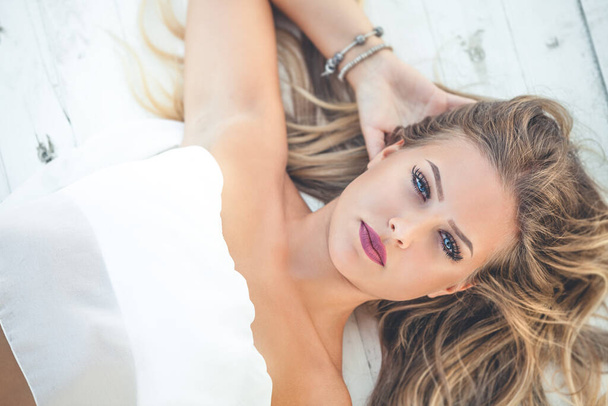 Beautiful young blonde woman with long hair lying on white wooden background. Blue eyes. The girl is relaxing and looks towards the camera. Summer lights. - Foto, Imagen
