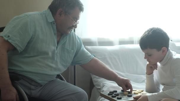 an elderly man in a wheelchair plays Board games with his grandson in the room. Nursing home. The boy takes care of an elderly grandfather. Caring for the disabled. - Séquence, vidéo