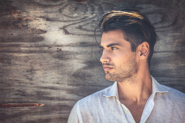 Profile portrait of young and handsome man on wooden background. He is wearing a white shirt and has a light beard. - Foto, Imagen