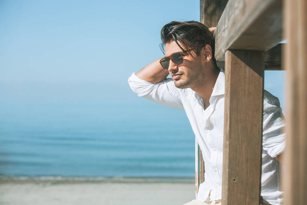 Fiatal, vonzó férfi napszemüvegben, aki a tengerre néz a nyár folyamán. Előre néz, fehér ingbe öltözik, és egy fából készült építkezésre támaszkodik.. - Fotó, kép