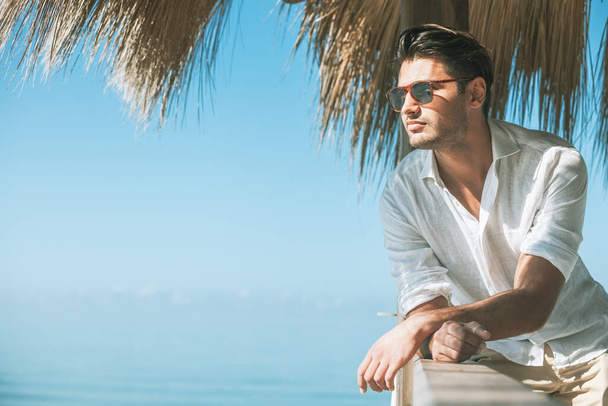 Giovane uomo attraente con occhiali da sole che si affaccia sul mare durante l'estate. Guardando avanti, vestito con una camicia bianca e appoggiato su una costruzione in legno
. - Foto, immagini