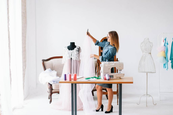 Attractive stylish seamstress or clothing designer measuring a length of neutral colored fabric alongside her sewing machine with copyspace - Photo, Image