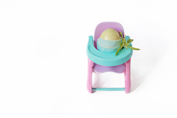 Πασχαλινό αυγό σε μια προστατευτική μάσκα κάθεται σε μια υπερυψωμένη καρέκλα με ένα μπουκέτο. Πασχαλινές διακοπές κατά τη διάρκεια της πανδημίας coronavirus. - Φωτογραφία, εικόνα