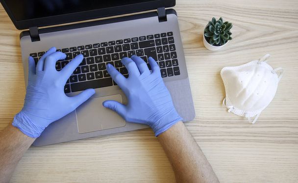 Männliche Hände in blauen Schutzhandschuhen arbeiten an einem modernen Laptop. Facharzt in blauen Latexhandschuhen. Auf dem Schreibtisch liegt eine medizinische Maske. Viren- und Bakterienschutzkonzept. Ansicht von oben. - Foto, Bild