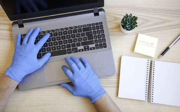Чоловічі руки в синіх захисних рукавичках працюють на сучасному ноутбуці. Спеціаліст з синіх латексних рукавичок. Медична маска на столі. Концепція захисту вірусів і бактерій. Концепція Covid-19
. - Фото, зображення