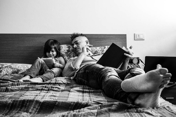 Vader en dochter in casual denim kleren zitten samen op bed. Het meisje dat plezier heeft met schrijven of tekenen terwijl de vader naar haar kijkt. Jeugd en ouderschap concept. Vrijetijdsbesteding thuis. - Foto, afbeelding