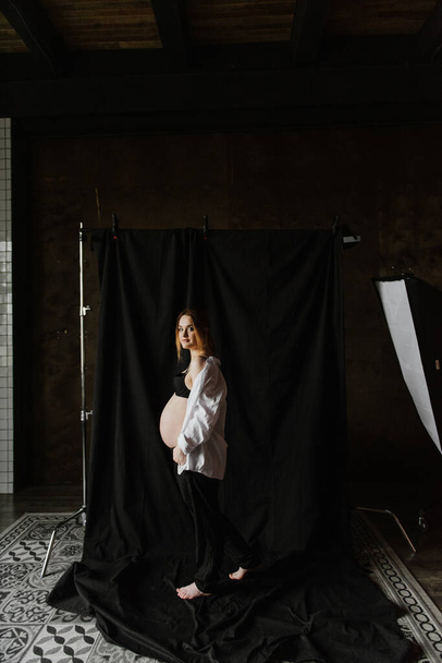 Young pregnant woman in underwear and a white shirt near fabric background in the studio. - Foto, Bild