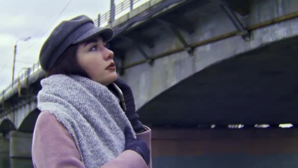 橋や川の背景に対するピンクのコートの若い女性の肖像画。ストック映像だ。暖かいキャップとスカーフを身に着けている静かで美しい女性は悲しいと情熱的に見えます. - 映像、動画