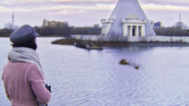 Jeune femme pensive en manteau rose debout au bord du lac regardant de côté, effet caméra VHS. Images d'archives. Jeune fille aux cheveux longs foncés debout près de l'eau en saison froide
. - Photo, image