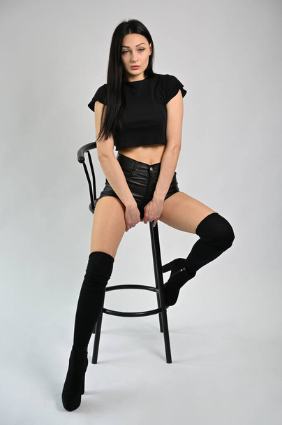 Studio photo of a beautiful and fashionable brunette model in glamorous clothes. Sympathetic girl is sitting on a chair. - Foto, Imagem