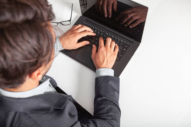 Hombre con computadora portátil escribiendo en el teclado. En la mesa blanca. El hombre lleva un traje y una camisa. Gafas graduadas apoyadas sobre la mesa
. - Foto, Imagen