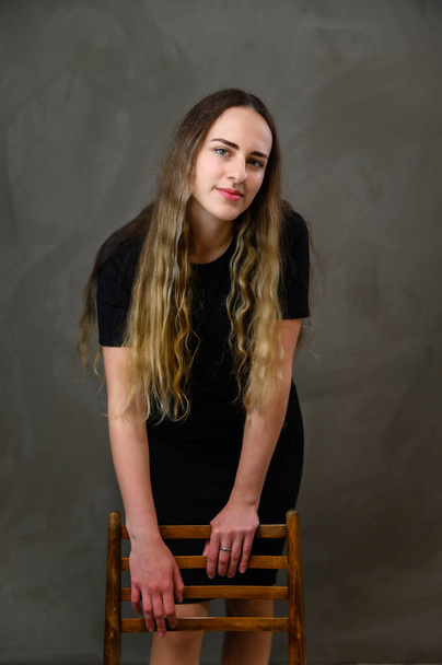 Vertical studio photo on a gray background. Cute Caucasian teenager girl in a black dress with long hair. A model stands next to a chair - Foto, imagen